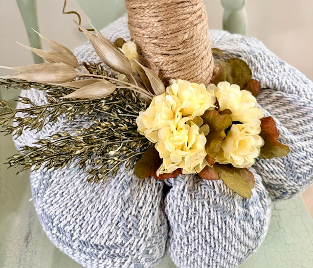 A decorative knitted pumpkin centerpiece with a gray and white marled knit body, a wrapped twine pole, pale yellow faux flowers, and dried botanical accents, shown close up on a pale green surface.