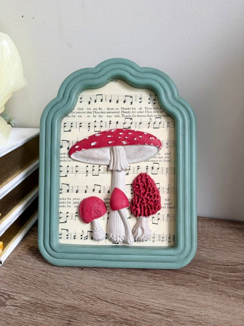 A decorative framed wall art piece featuring four red and white mushrooms on a sheet music background, set in a scalloped mint green frame on a wooden surface.