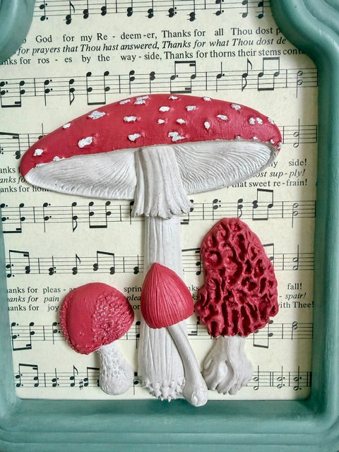 Three-dimensional mushroom wall decor featuring a large red and white toad mushroom, a small red and white mushroom, and a textured red bracket mushroom, mounted on a vintage sheet music background with a teal frame visible.