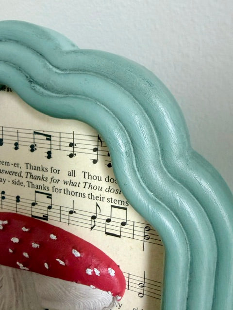 A close-up of a decorative wavy frame in a light mint or seafoam green color, holding vintage sheet music with a red and white spotted mushroom illustration.