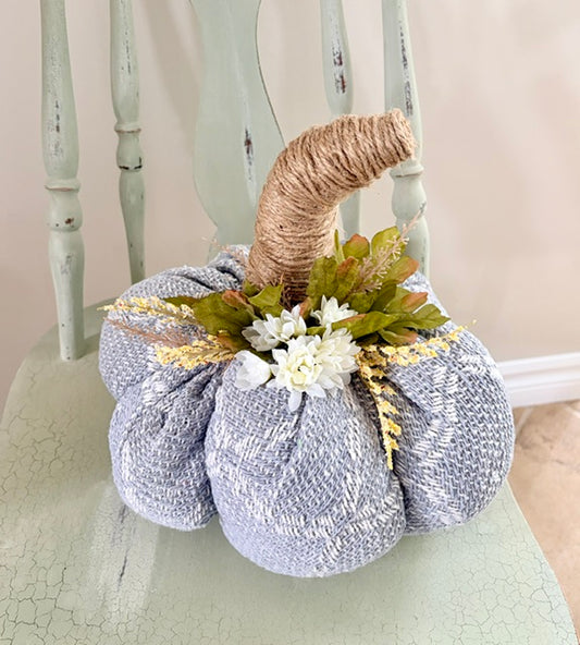 A decorative fabric pumpkin in a textured light blue-gray material, wrapped with natural jute twine at the stem, and topped with faux white flowers and green and gold leaves, displayed on a distressed mint green chair.