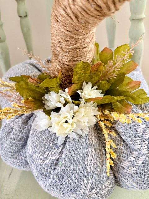 A knitted decorative pumpkin in heathered light gray, wrapped with natural jute twine at the top and adorned with faux white flowers, green leaves, and yellow dried stems, displayed on a painted chair.