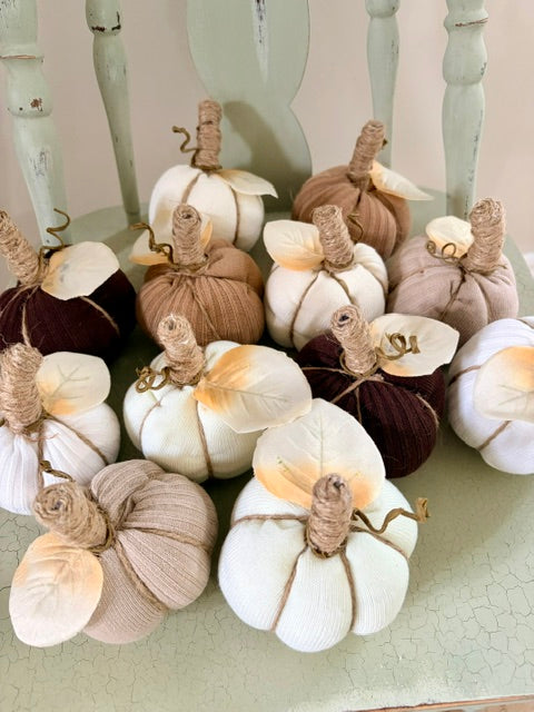A group of decorative knitted pumpkins in neutral fall colors, including cream, beige, taupe, and dark brown, arranged on a painted wooden chair.