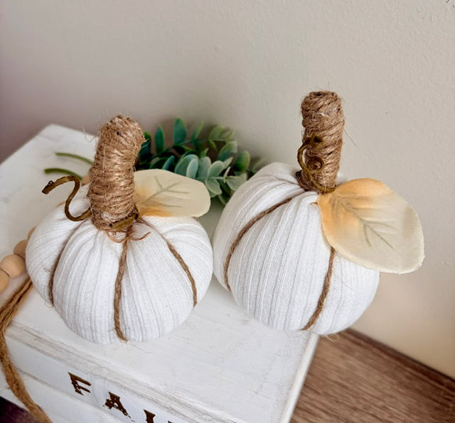 Two white fabric pumpkins with brown twine stems and pale leaves, displayed on a white wooden box with a small green plant in the background.