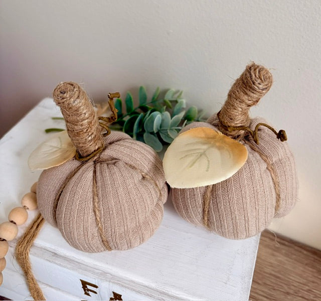 Two decorative fabric pumpkins in a warm beige color, wrapped with natural jute stems and cream-colored leaves, displayed on a white wooden box with a wooden bead garland.