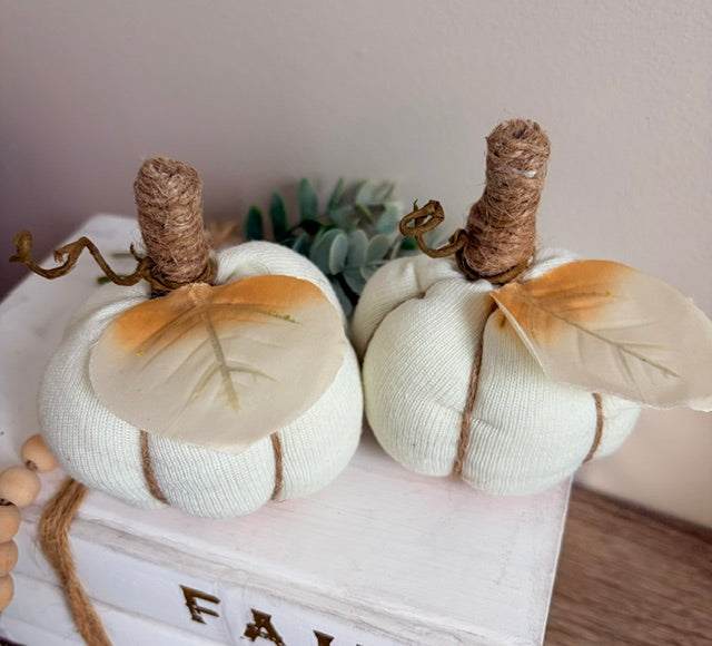 Two small fabric pumpkins in a warm off-white color, wrapped with natural jute twigs and decorated with dried orange and beige leaves, displayed on a white wooden box.