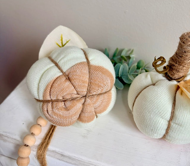 Two fabric decorative pumpkins styled on a white wooden surface. The larger pumpkin on the left is off-white with a peach-pink center, wrapped in twine and tied with a bow, with a wooden bead garland nearby.