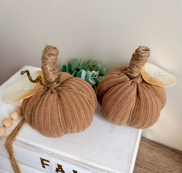 Two decorative fabric pumpkins in a warm tan-brown color, styled on a white wooden box with a small succulent and wooden beads in the background.