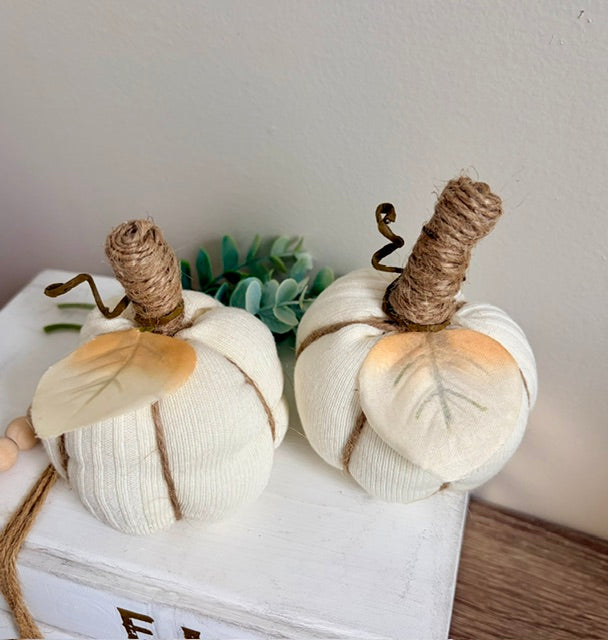 Two cream knit fabric pumpkins with natural jute-wrapped stems and painted orange leaf accents, displayed on a white wooden box with a small green plant in the background.