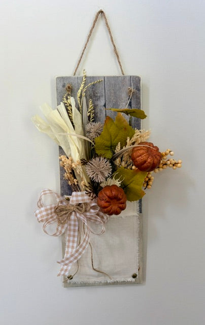 A rustic autumn-themed wall hanging featuring a weathered gray wooden plaque with a fabric pocket, decorated with faux dried grasses, two small orange pumpkins, green leaves, and a beige-and-white gingham ribbon bow, suspended by a twine hanger against a plain white wall.