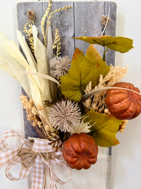 Autumn-themed decorative wall hanging featuring faux dried grasses, olive and mustard leaves, two small copper-orange pumpkins, and a beige gingham ribbon bow, mounted on a weathered gray wooden panel.
