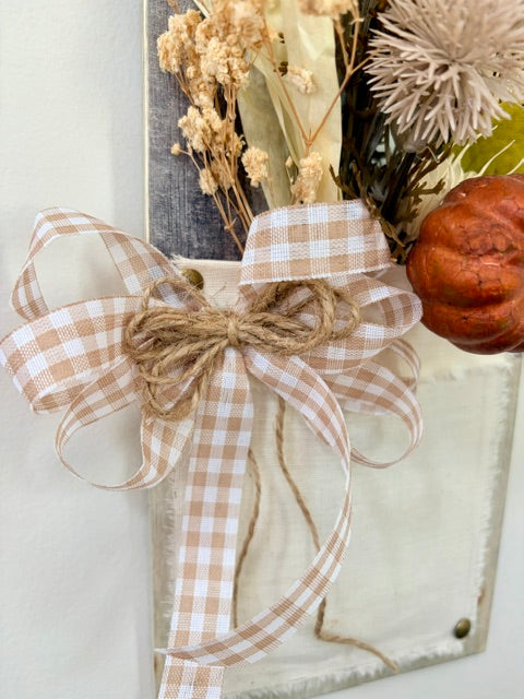A close-up of a rustic autumn decorative arrangement featuring a large beige and white gingham ribbon bow tied with twine, dried flowers, a small orange pumpkin, and a weathered white wooden panel against a light wall.