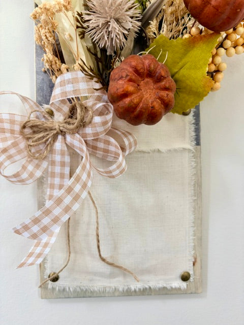 Autumn-themed rustic wall hanging featuring a faux linen panel with a large beige-and-white gingham ribbon bow, dried flowers, small orange pumpkins, and green leaves.