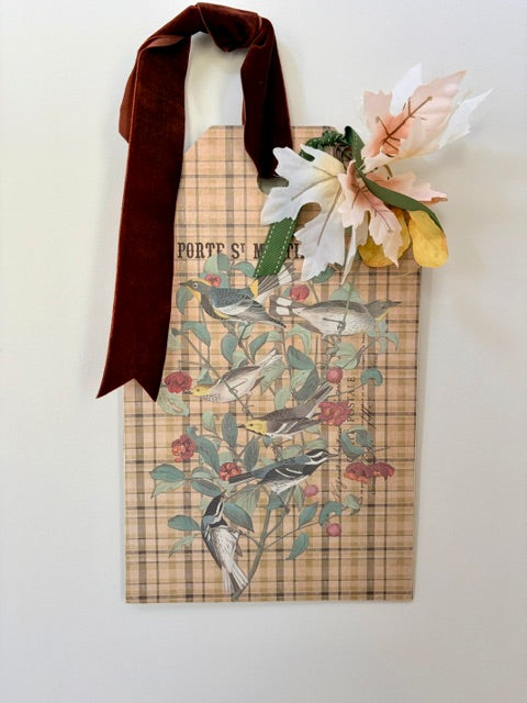 A decorative hanging wall sign featuring a vintage-style bird illustration over a beige plaid background, with a dark brown velvet ribbon handle and faux white and yellow flowers at the top, shown against a plain white wall.