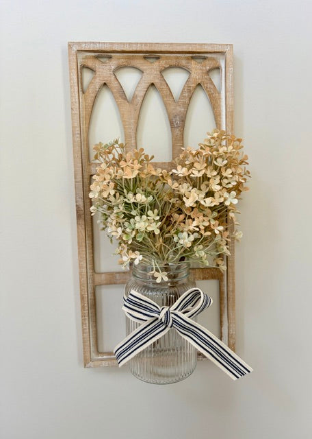 A rustic wooden wall hanging with an arched cutout design, holding a clear ribbed glass jar vase filled with dried cream and tan flowers, tied with a navy and cream striped ribbon bow, against a plain light wall.