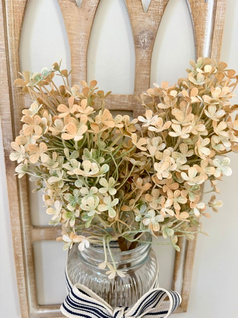 A decorative arrangement of dried pale peach and cream flowers with muted green stems, displayed in a clear ribbed glass jar tied with a navy-and-white striped ribbon, set against a distressed wood panel background.