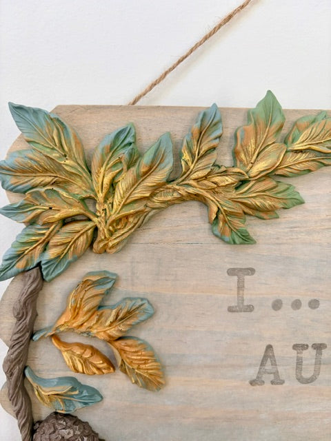 A rustic wooden wall hanging or plaque with embossed leafy branches in green and gold tones, hanging from a rope against a plain light background. The wood shows partial engraved text reading 'I .