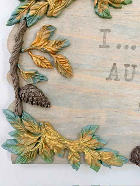 Close-up of a decorative wooden sign with raised autumn leaves in green and gold, a pinecone, and partial stamped text reading 'I.