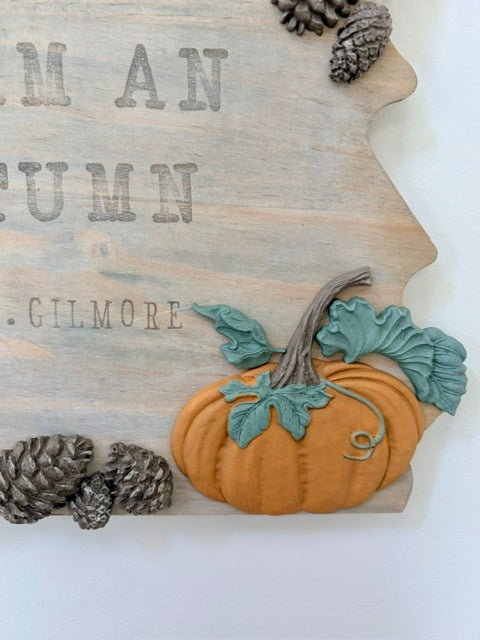 A rustic autumn-themed wooden sign shaped like the state of Georgia, featuring a raised orange pumpkin with teal leaves, pinecones, and partial text reading 'AN SUMM .GILMORE' on a white background.