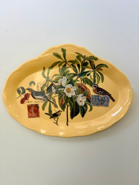 A yellow ceramic serving platter featuring a vintage-style botanical illustration with green leaves, white flowers, birds, and postage stamp motifs, shown on a plain light background.