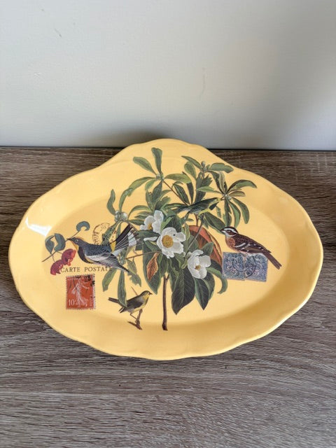 A yellow ceramic serving platter with a scalloped rim, decorated with a botanical and bird illustration featuring white flowers, green leaves, and birds, resting on a gray wooden surface.