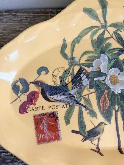 A close-up of a decorative ceramic plate with a pale yellow background, featuring painted birds, white flowers, green leaves, and a red postage stamp illustration with the text 'CARTE POSTALE'.