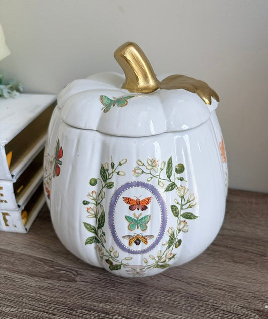 A white ceramic pumpkin-shaped lidded container with a gold-painted stem, decorated with colorful butterflies and floral motifs, sitting on a wooden surface.