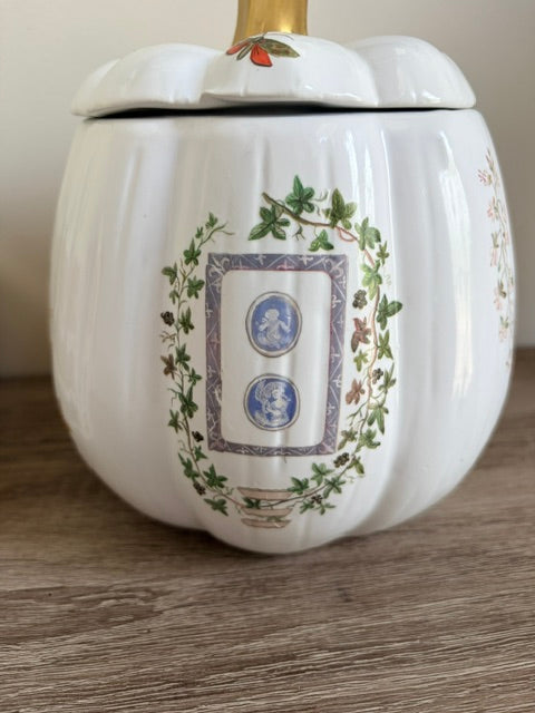 A white ceramic pumpkin-shaped jar with a lid, decorated with green ivy, blue cameo-style portraits, and a gold-toned handle, shown on a wooden surface.