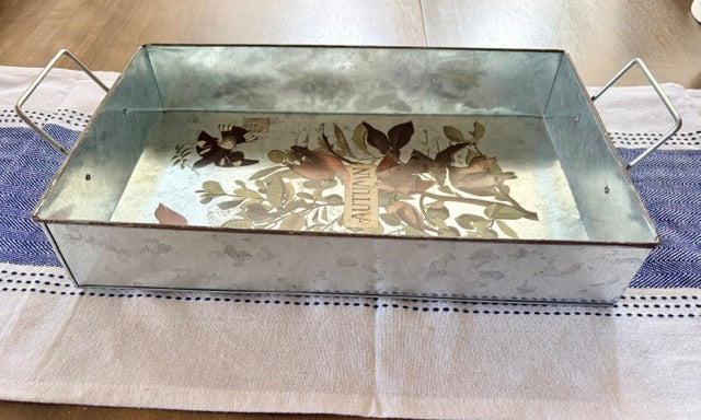 A rectangular galvanized metal tray with wire handles, featuring a vintage-style floral and bird illustration printed inside, resting on a white tablecloth with blue stripes.