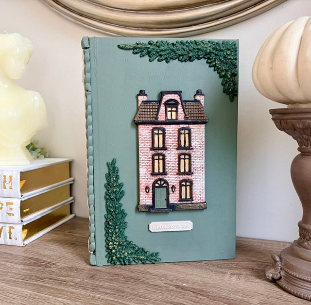 A decorative hardcover notebook or journal with a teal-green cover, featuring an embossed pink house illustration and green tree accents, displayed on a wooden surface next to decorative objects.
