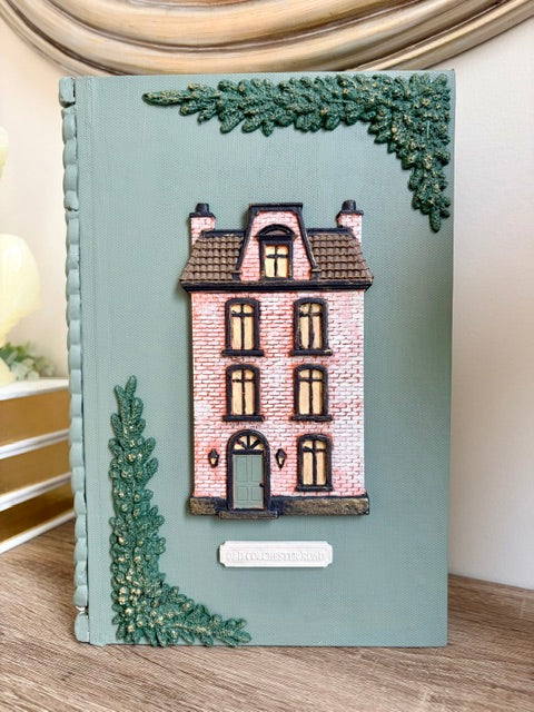 A book box featuring a 3D pink brick house with a green roof, decorated with textured green foliage in the corners on a sage green background.
