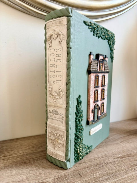 A decorative book-shaped object with a pale green cover and spine, featuring embossed ornamental detailing, a small brick house illustration on the front, and green vine accents, standing on a wooden surface against a light wall.