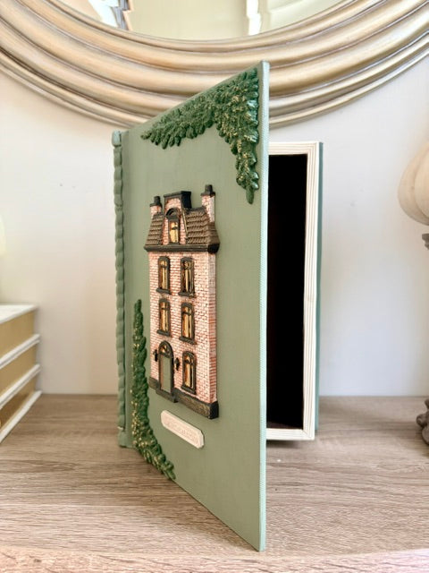 A decorative hardcover notebook or journal with a sage green cover, featuring a 3D embossed house design and green leafy accents, standing open on a wooden surface.