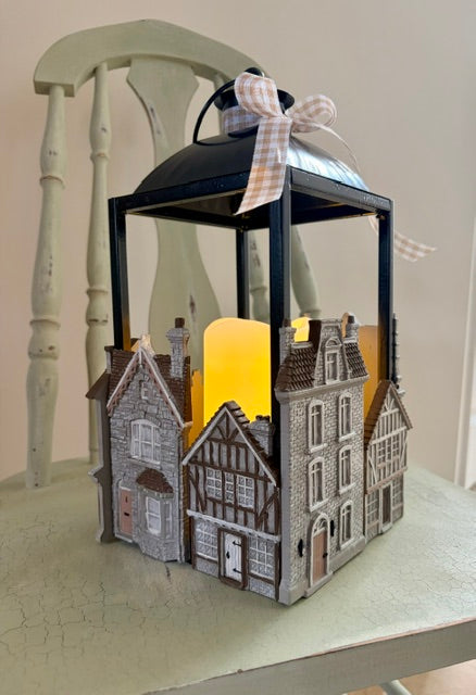 A decorative lantern shaped like a row of old European-style houses, with a glowing yellow candle inside and a black metal frame topped with a silver lid and a gingham ribbon bow. The lantern sits on a distressed light green table in front of a matching chair.