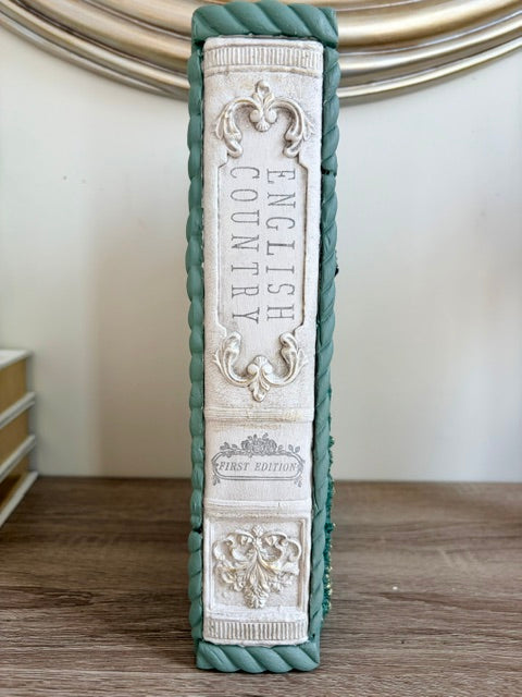 A decorative book spine prop featuring 'ENGLISH COUNTRY' and 'FIRST EDITION' in raised white embossed lettering, bordered by a twisted teal-green frame, standing on a wooden surface.