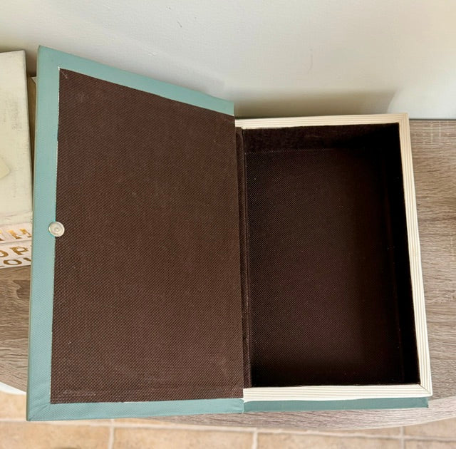 An open book-shaped storage box with a teal-green cover, dark brown fabric-textured inner panels, and a light wood frame, photographed on a wood surface against a white wall.