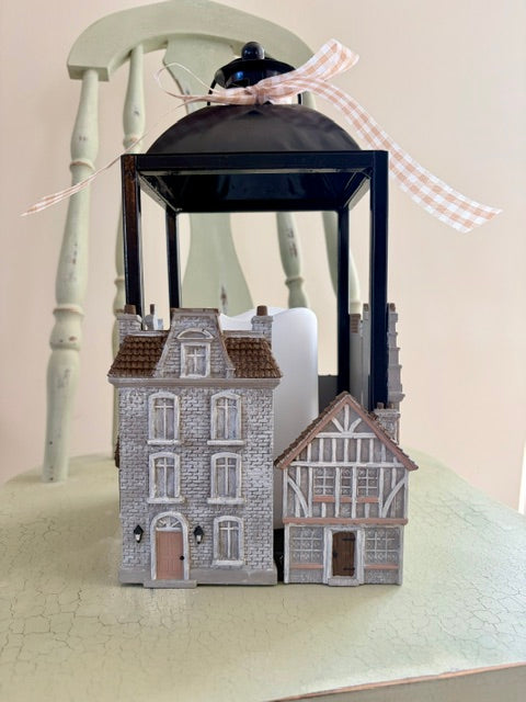 A decorative black metal lantern with a small black bird figurine on top, decorated with a pink-and-white gingham ribbon. In front of the lantern are two miniature house models, one gray stone-textured and one brown-and-beige timber-framed, set on a light green surface.