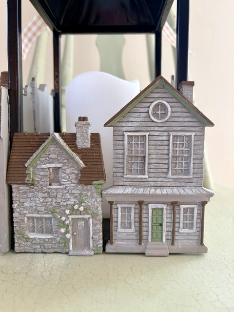 Two miniature decorative houses displayed side by side on a light surface under a black shelf or frame. The left house is small with a gray stone facade, brown roof, and white trim.