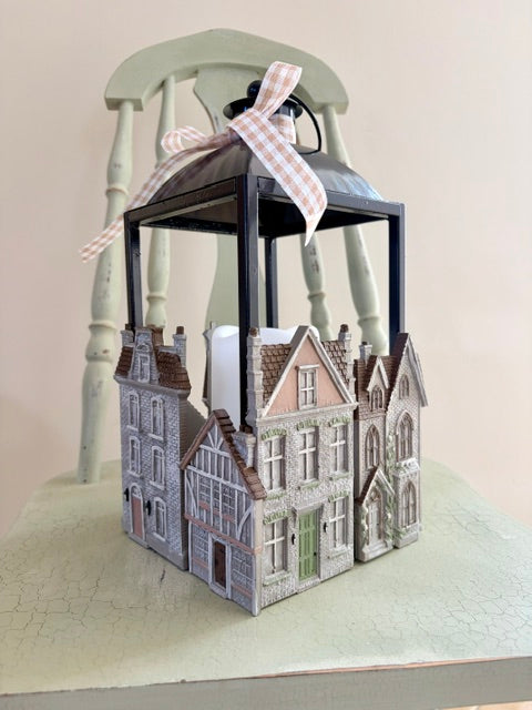 A decorative miniature house-shaped lantern with a black metal frame and a pink-and-white gingham ribbon bow on top, displayed against a pale green chair and cream background.
