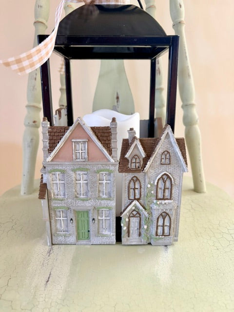 Two miniature Victorian-style house figurines with detailed windows, brown roofs, and painted brick textures, displayed in front of a black lantern on a light-colored surface.