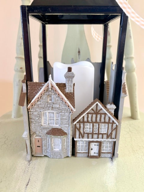 A decorative miniature village house set displayed inside a black lantern, featuring two detailed cottages with gray stone and timber facades, white candles, and a soft beige background.