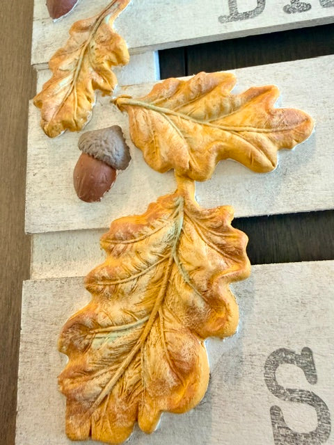 Acrylic wall decor featuring textured golden-brown oak leaves and acorns mounted on a rustic light wood plank background with dark lettering.