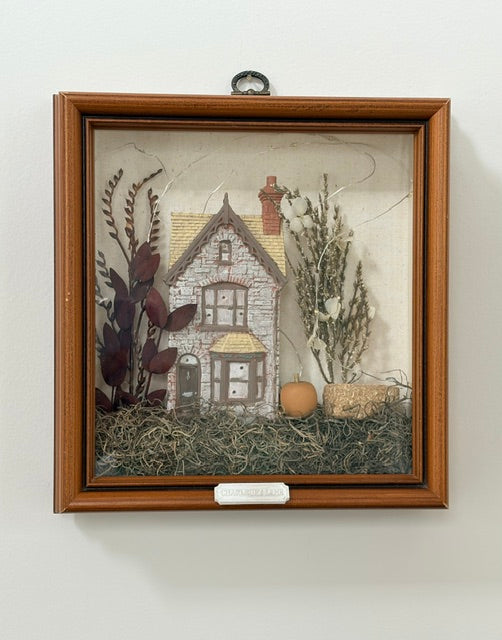 A framed decorative shadow box featuring a small house surrounded by dried plants and grass, mounted on a light wall.