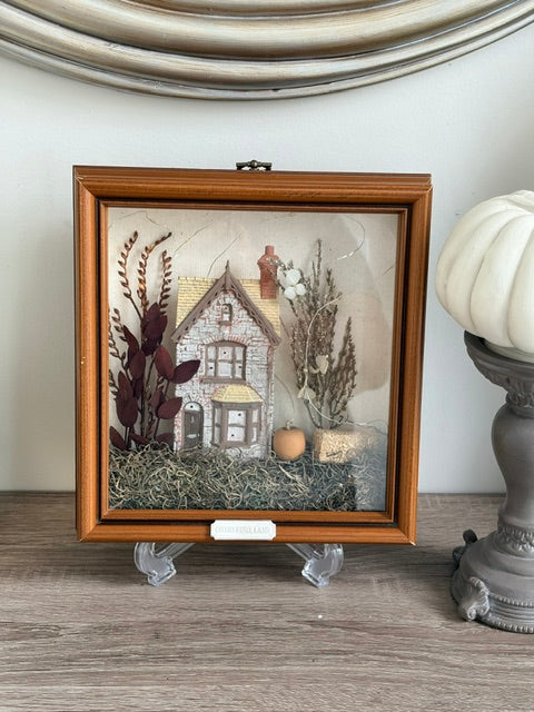 A square wooden shadow box display featuring a miniature house scene with dried flowers, grass, and small decorative elements under clear glass, shown on a tabletop with a white pumpkin and gray pedestal beside it.