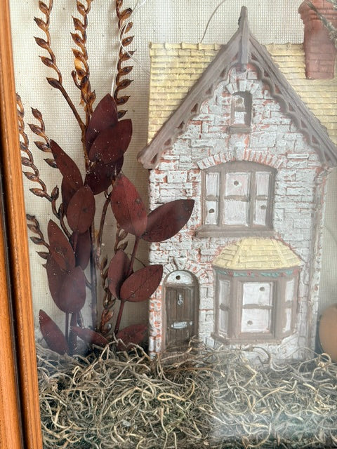A decorative miniature house ornament or wall hanging with a gray and white brick-textured facade, yellow shingled roof, small bay window, and a doorway, displayed with dried brown leaves and straw-like material against a neutral fabric background.