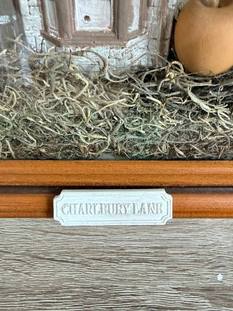 A close-up of a miniature decorative scene featuring a faux wood door, a brown ceramic pumpkin, and a layer of dried moss or pine needles. A small white plaque in the foreground reads 'CHARLBURY LANE'.
