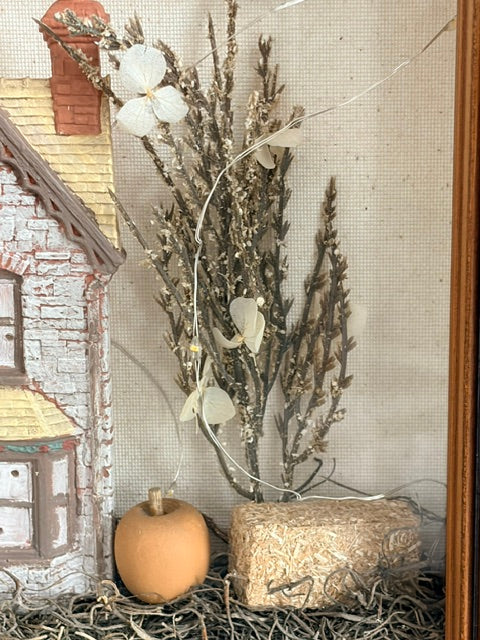 A rustic autumn-themed decorative display featuring dried botanical stems, white flower petals, a small orange pumpkin, a textured hay bale, and a miniature house on a straw base against a neutral fabric backdrop.
