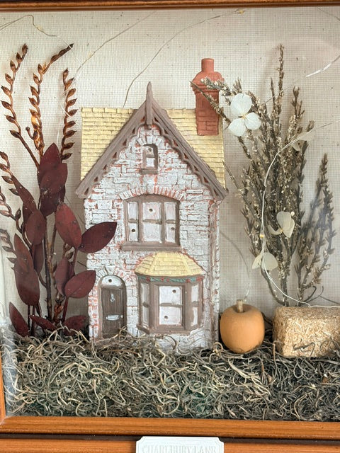 A framed decorative display featuring a small hand-painted cottage surrounded by dried botanicals, a small orange pumpkin, and hay, set against a neutral textured background.