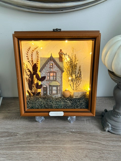 A square wooden shadow box display featuring a miniature house scene with warm string lights, dried botanical elements, and a small pumpkin, shown on a tabletop indoors.