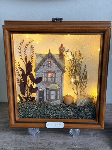 A framed shadow box display featuring a miniature stone house, dried botanical elements, warm string lights, and a small pumpkin inside a wooden frame, shown on a tabletop against a plain wall.